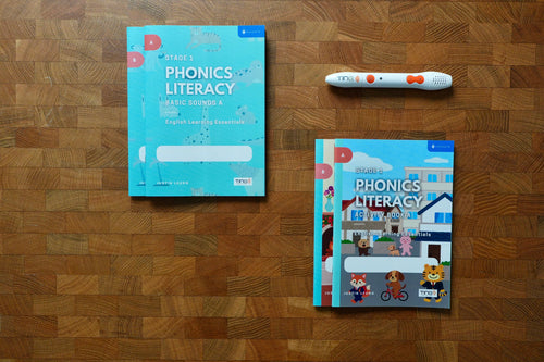 A phonics literacy learning kit arranged on a wooden surface, including teal workbook covers, a colourful children's key readers book, and a white tracking pen with orange accents.