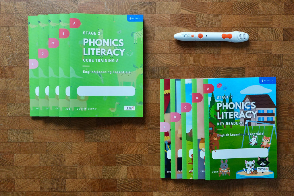 A phonics literacy learning kit arranged on a wooden surface, including green workbook covers, a colorful children's key readers book, and a white tracking pen with orange accents.