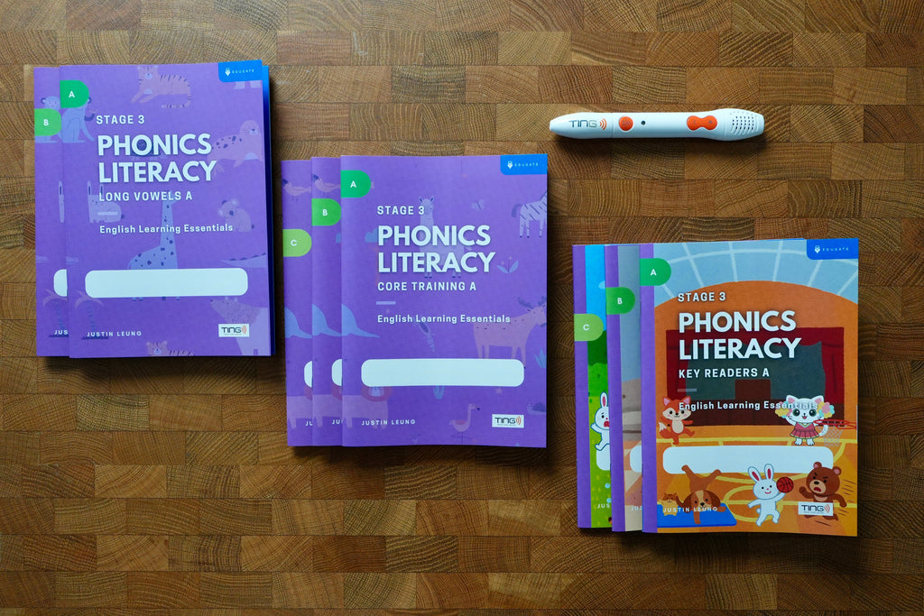 A phonics literacy learning kit arranged on a wooden surface, including purple workbook covers, a colorful children's key readers book, and a white tracking pen with orange accents.

