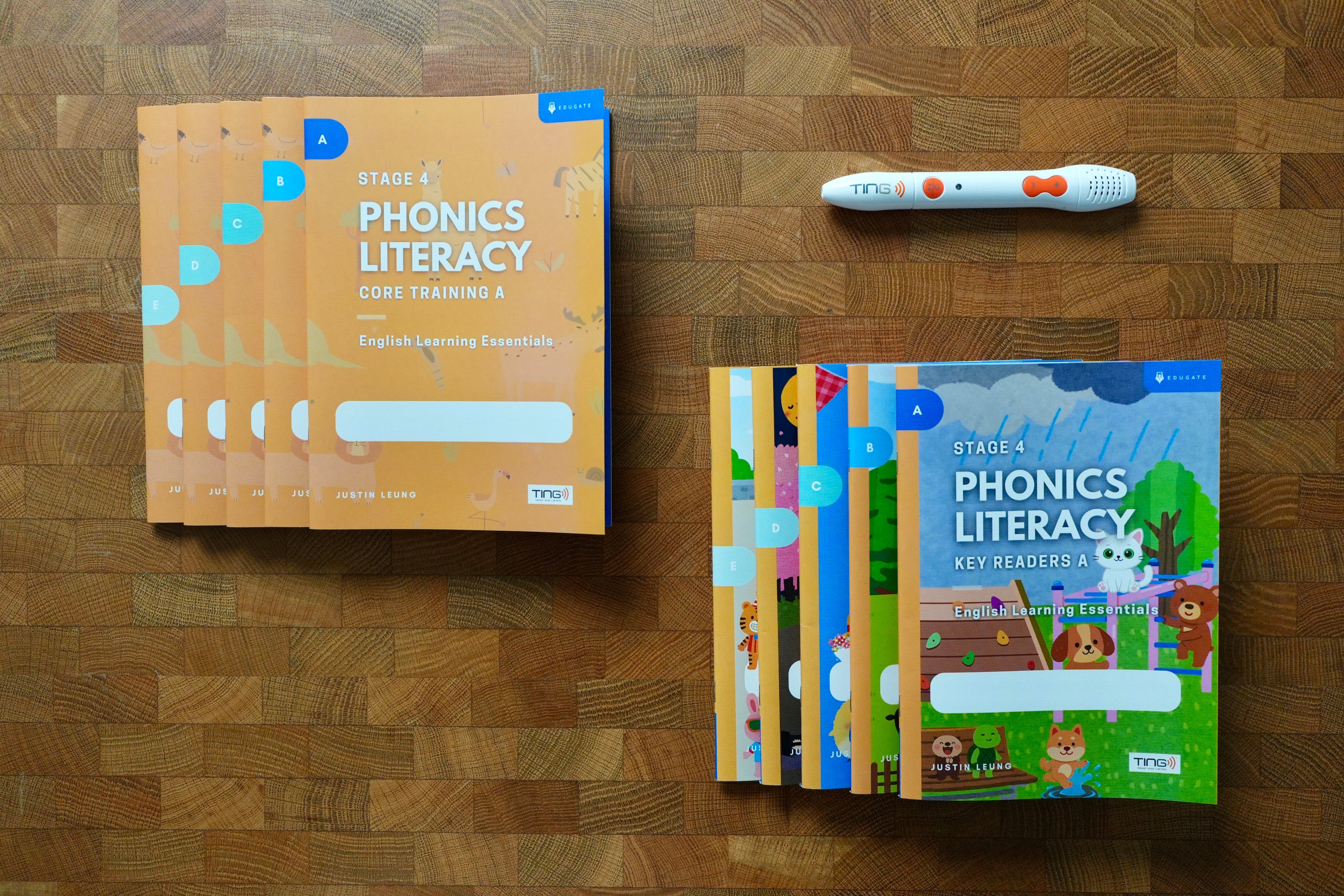 A phonics literacy learning kit arranged on a wooden surface, including orange workbook covers, a colourful children's key readers book, and a white tracking pen with orange accents.