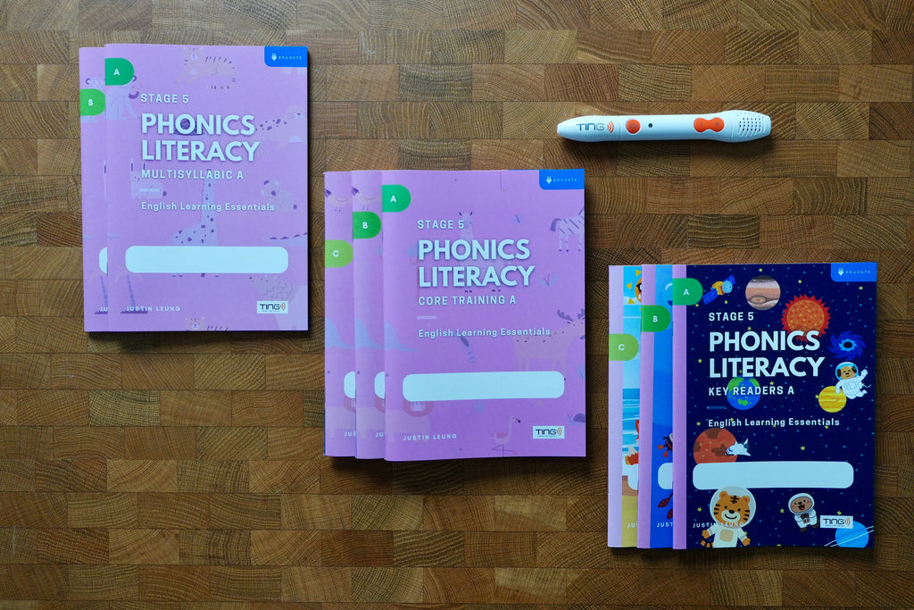 A phonics literacy learning kit arranged on a wooden surface, including pink workbook covers, a colorful children's key readers book, and a white tracking pen with orange accents.