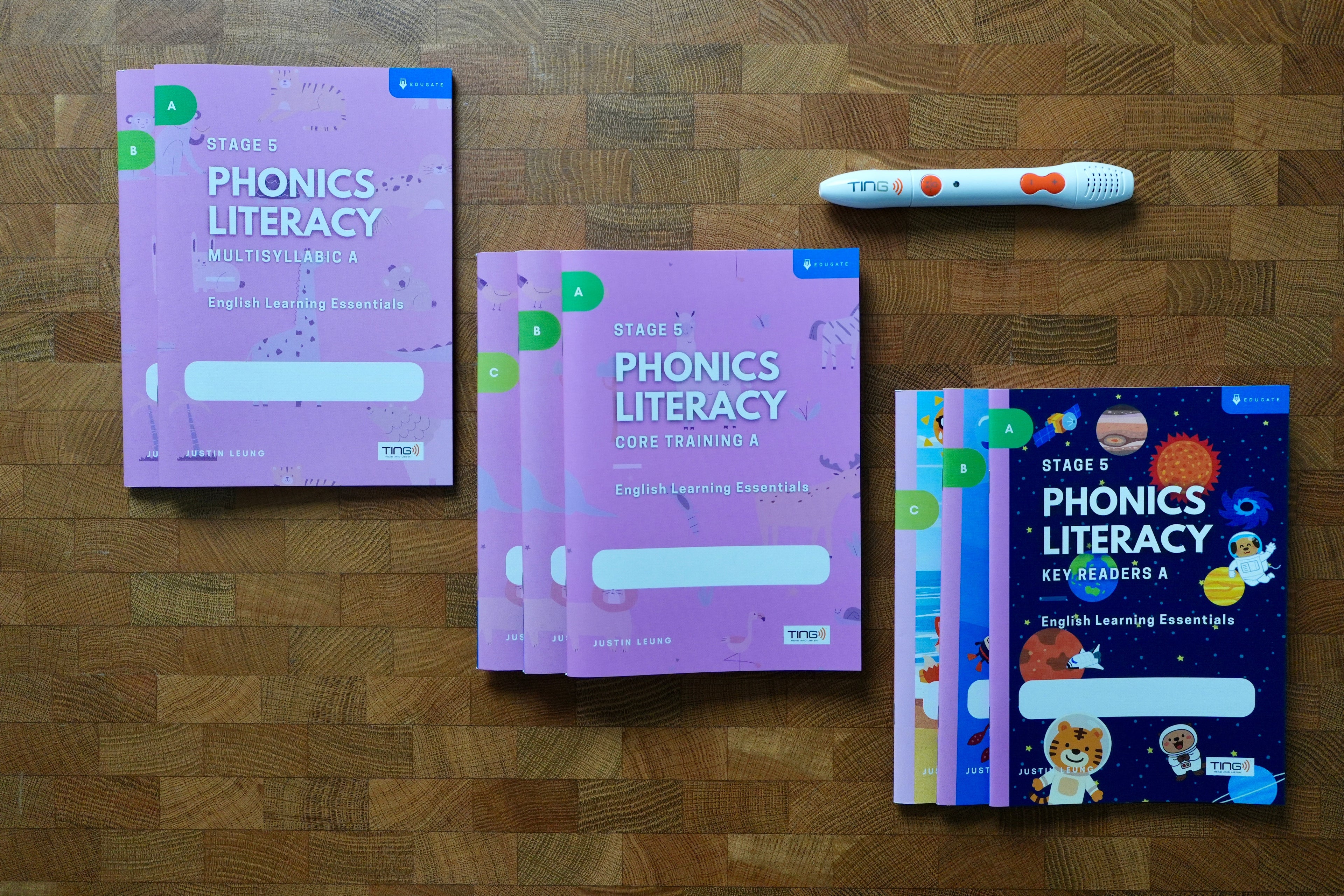 A phonics literacy learning kit arranged on a wooden surface, including pink workbook covers, a colorful children's key readers book, and a white tracking pen with orange accents.