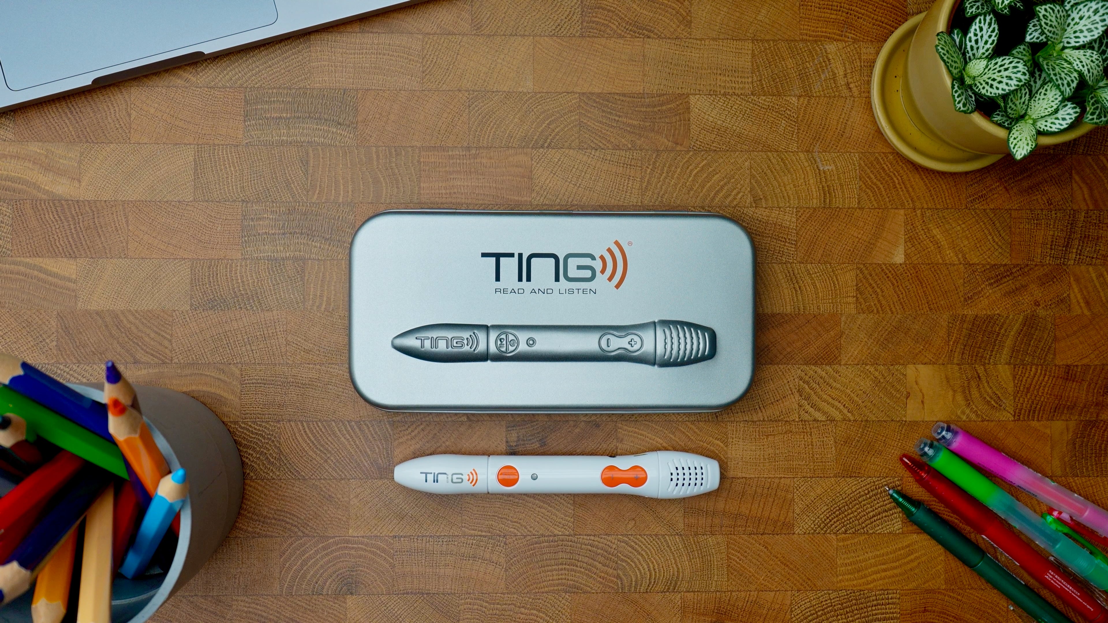 A TING Read and Listen tablet and its charging case are arranged on a wooden desk with school supplies and a potted plant nearby.