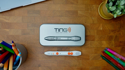 A TING Read and Listen tablet and its charging case are arranged on a wooden desk with school supplies and a potted plant nearby.